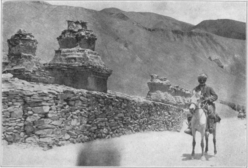 A man in a turban riding a horse alongside a stone wall, with two small structures and mountains in the background.
