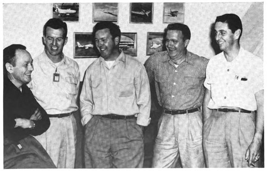 One man sitting and five men standing, all looking happy and relaxed