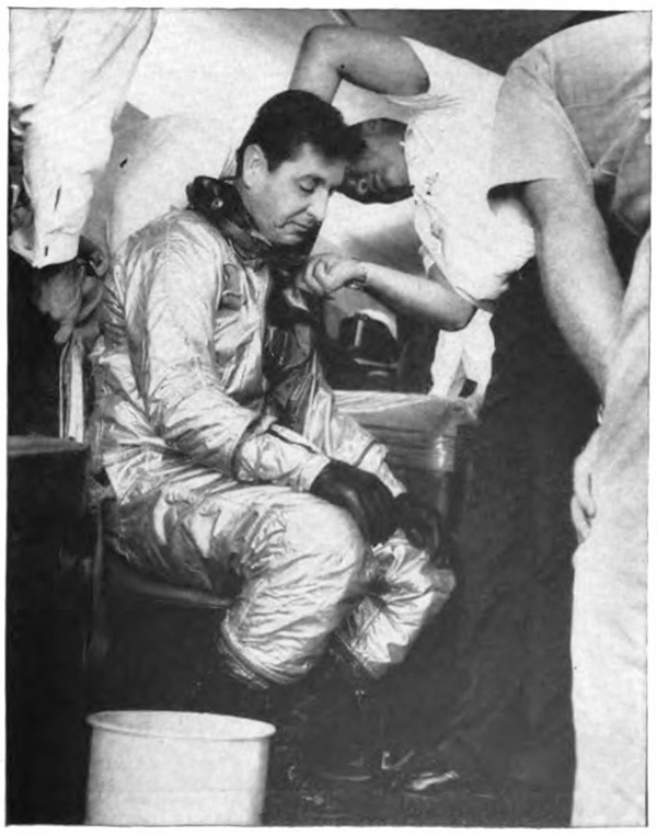 A man sitting in a protective suit while another man inspects the suit