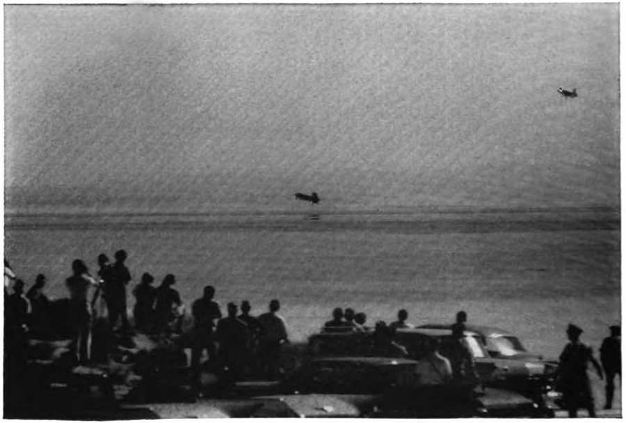 A crowd of people watching two planes from a lakeshore; some are standing on cars