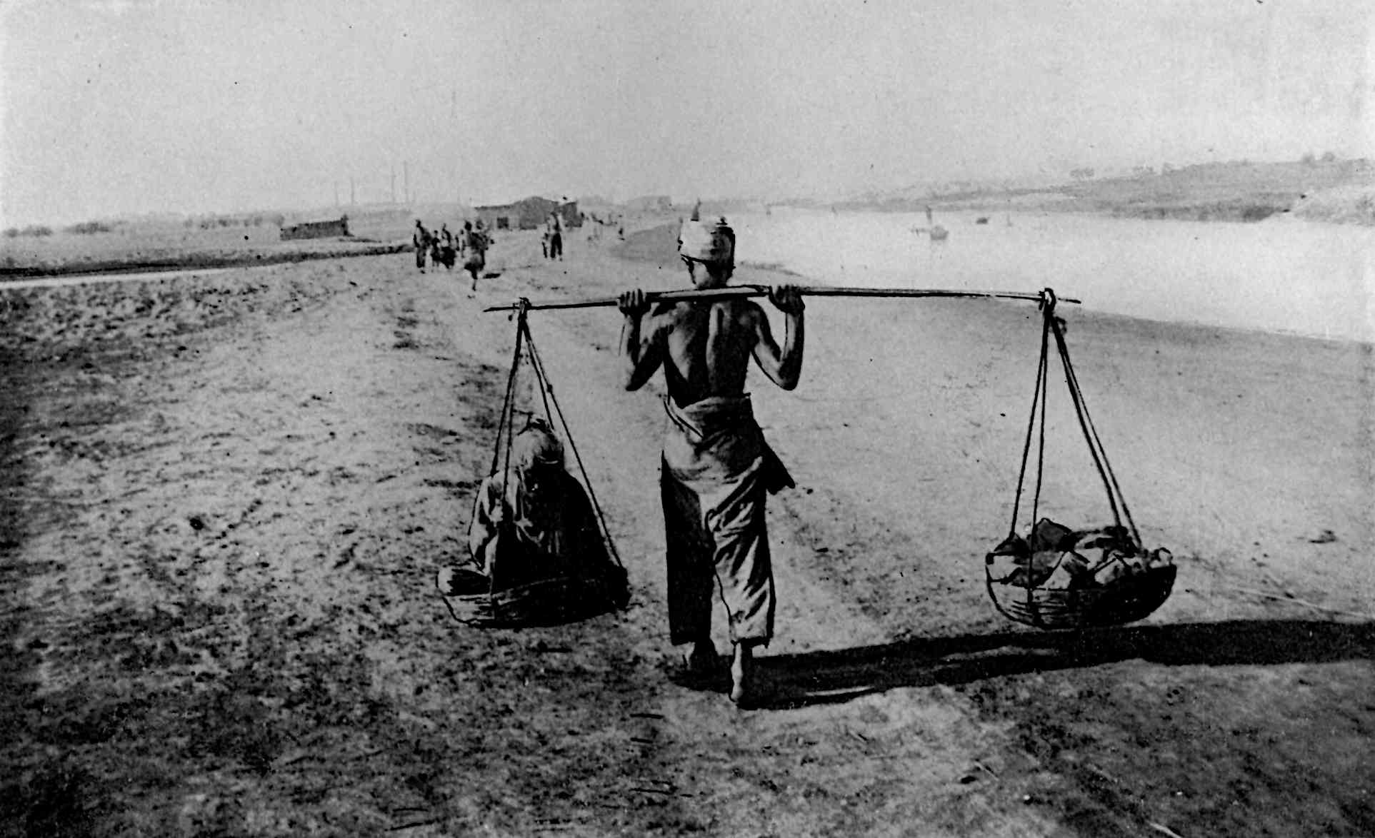 A man carrying 2 bowls of items on a stick on the shoulders