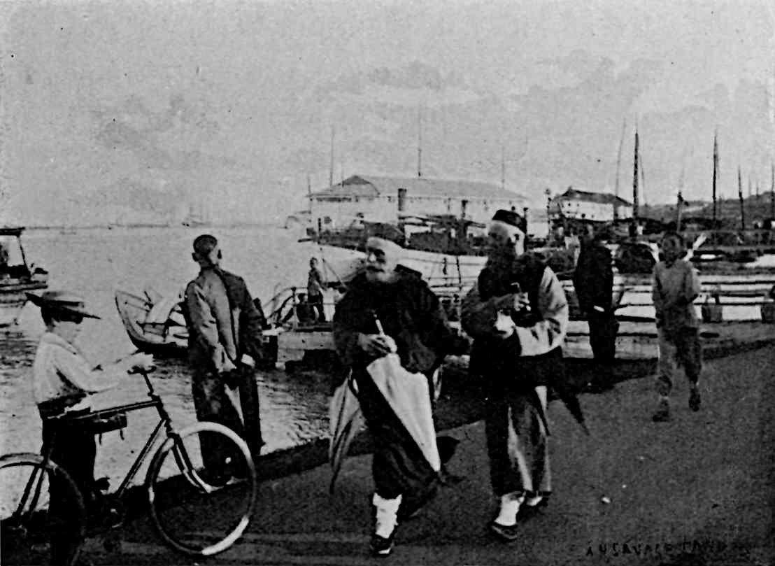2 bearded Western men in unique attire walking on a harbor with boats, a man appears to laugh behind them