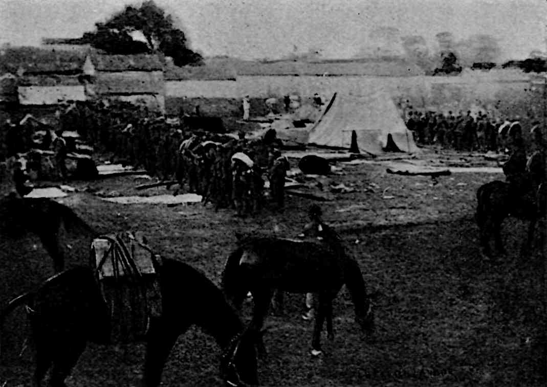 People in uniform in line, with horses in the front and tents and buildings in the back